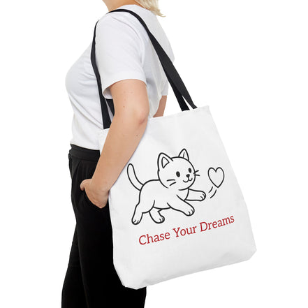 Person holding a white tote bag with a cat illustration and 'Chase Your Dreams' text.