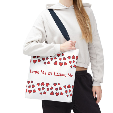 Person holding a tote bag with red hearts and text, wearing a light gray hoodie and black pants.