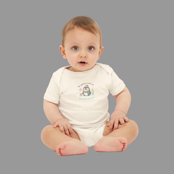 Penguin baby onesie Be Patient With Me This is My First Christmas infant bodysuit. Baby sitting on a white background wearing a white onesie with a penguin design.