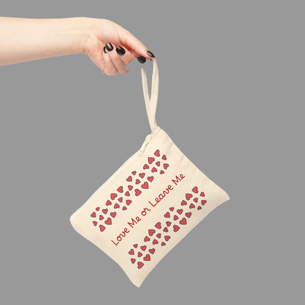 Love Me or Leave Me zipper pouch with romantic heart pattern cosmetic bag accessory. Beige pouch with red heart patterns and text held by a hand on a gray background