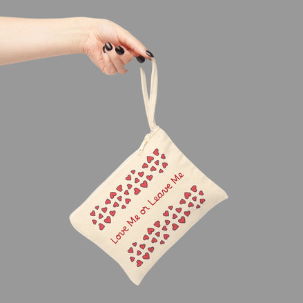Love Me or Leave Me zipper pouch with romantic heart pattern cosmetic bag accessory. Beige pouch with red heart patterns and text held by a hand on a gray background